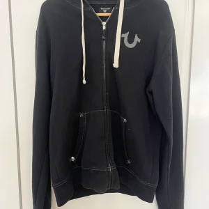 Svart hoodie från True Religion - Svart hoodie med dragkedja från True Religion. Tröjan har vit logga på bröstet och stort tryck på ryggen. Den är i bra skick, inga flaws! 