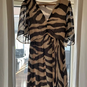 Zebramönstrad klänning från H&M - Säljer en luftig zebramönstrad klänning från H&M med v-ringning och korta, vida ärmar. Klänningen har svart och beige mönster och är tillverkad i ett lätt, syntetiskt material. Perfekt för dig som gillar djurmönster och vill sticka ut.