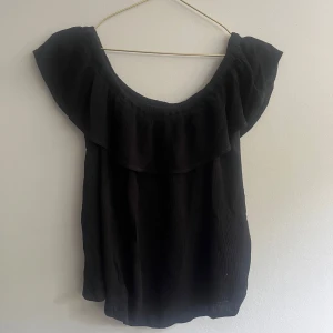 Svart volangtopp från ICHI - Svart topp från ICHI med bred volang runt halsringningen. Toppen är ärmlös och har en luftig, lätt struktur. Perfekt för varma dagar och enkel att matcha med olika stilar.