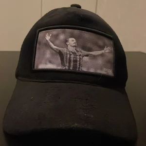 Svart keps med fotbollstryck från BOZZ - Svart keps med böjd skärm och mesh på sidorna. Framtill finns ett svartvitt tryck av en fotbollsspelare med utsträckta armar. Märket BOZZ syns på framsidan. Justerbar passform baktill.