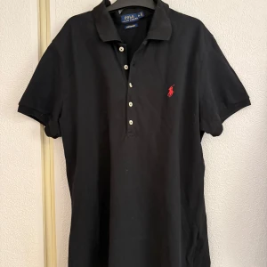 Svart pikétröja från Polo Ralph Lauren - Klassisk svart pikétröja från Polo Ralph Lauren med korta ärmar och knappar framtill. Tröjan har den ikoniska röda loggan broderad på bröstet och är tillverkad i bomullsmaterial med stretch mesh för extra komfort.