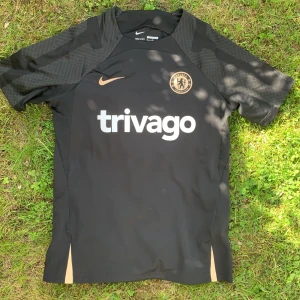 Chelsea svart tränings t-shirt från Nike - Chelsea FC svart tränings t-shirt från Nike med guldiga detaljer och klubbmärke på bröstet. Tröjan har korta ärmar, Dri-Fit material och slim fit passform. Stor trivago-logga på framsidan.