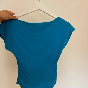Turkos offshoulder topp - Säljer en turkos offshoulder topp med korta ärmar och öppen rygg. Toppens material är mjukt och stretchigt, perfekt för varma dagar. Snygg och enkel design som passar till många olika outfits.