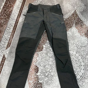 Grå och svarta friluftsbyxor från Lundhags - Säljer ett par slitstarka friluftsbyxor från Lundhags i grått och svart. Byxorna har flera fickor med dragkedja och förstärkta partier på knän och benslut. Perfekta för utomhusaktiviteter och äventyr.