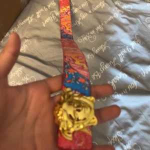 Versace bälte - Säljer ett unikt bälte med färgglatt mönster i rött, blått och orange samt ett stort guldfärgat lejonspänne. Bältet har en konstnärlig design och insidan är ljusbeige. Perfekt för dig som vill sticka ut med accessoarer.