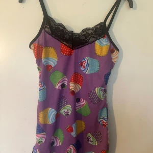 Lila linne med cupcakes och spets S/M fr Insanity - Säljer ett färgglatt lila linne med cupcakes-mönster och svart spets upptill. Linnet har tunna axelband och är perfekt för dig som gillar roliga och unika plagg. Helt nytt. Lite stretchigt.