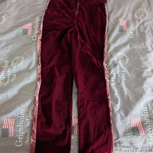 Vinröda sammetsbyxor med rosa sammetsband från NA-KD - Säljer ett par vinröda sammetsbyxor från NA-KD med breda ben och rosa sammetsband längs sidorna. Byxorna har normal passform. Perfekta för dig som vill ha något mjukt och snyggt.