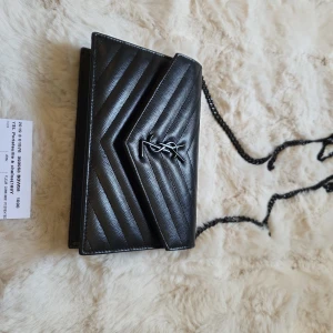 Svart axelväska från Saint Laurent - Säljer en svart axelväska från Saint Laurent med kedjerem och klassisk YSL-logga i silver på framsidan. Väskan är tillverkad i skinn med ett elegant, quiltat mönster i chevron-stil. Perfekt för dig som vill ha en stilren och lyxig accessoar.