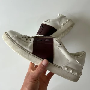 Valentino Garavani Open sneakers vit/vinröd - Snygga Valentino Garavani Open sneakers i vitt skinn med bred vinröd läderdetalj över snörningen. Platt sula med ikoniska nitar baktill och rund tå. Perfekta för dig som vill ha en lyxig och stilren look med en twist. Skriv för mer information🙌