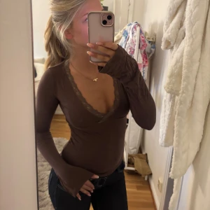 Brun långärmad topp med spets - Säljer en brun långärmad topp med djup v-ringning och fina spetsdetaljer längs urringningen. Modellen är tight och sitter snyggt på kroppen, perfekt att styla med jeans eller kjol. Materialet känns mjukt och stretchigt.