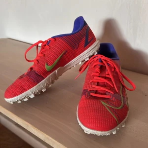 Nike fotbollsskor - Mercurial - Säljer ett par röda Nike fotbollsskor med gröna och blå detaljer. Skorna har snörning, mönstrad ovandel och vit sula med dobbar för bra grepp. Perfekta för spel på konstgräs eller grus. 
