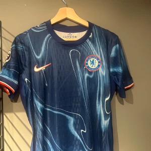 Chelsea FC blå matchtröja Nike - Chelsea FCs officiella matchtröja i mörkblått med unikt abstrakt mönster i ljusblått och vitt. Kortärmad modell med klubbmärke och Nike-logga på bröstet samt Premier League-märke på ärmen. Tillverkad i lätt och ventilerande material. Helt ny med tagg en replika i S