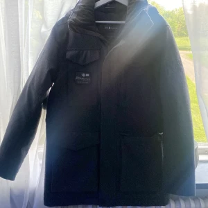 Svart parkajacka från Sail Racing - Svart parkajacka från Sail Racing med hög krage och flera praktiska fickor framtill. Jackan har en stilren look med diskret logga på bröstet och vänster ärm. Perfekt för kalla dagar med sin långa passform och rejäla huva.