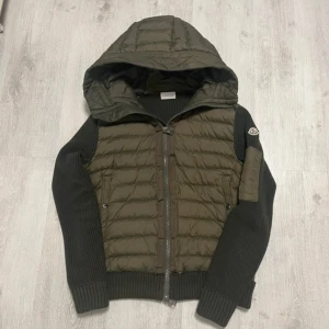 Moncler cardigan olive  - Äkta grön cardigan från Moncler med stickade ärmar och vadderad kropp.                                                     Jackan har huva, dragkedja framtill. Perfekt för höst och sommar, riktigt clean och modern look.                                       Priset är inte hugget i sten. Scanner på logga och QR kod du fungerar
