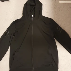 Svart hoodiejacka med dragkedja - Svart hoodiejacka med hel dragkedja framtill och huva. Jackan har ribbade muddar vid ärmslut och nederkant samt diskreta svarta ränder på ärmarna. Perfekt för en avslappnad streetstyle-look.