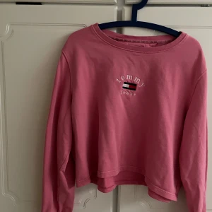 Rosa croppad sweatshirt Tommy Jeans - Säljer en rosa croppad sweatshirt från Tommy Jeans med broderad logga och flagga framtill. Tröjan har rund halsringning och långa ärmar. Perfekt för dig som gillar en avslappnad och trendig stil.