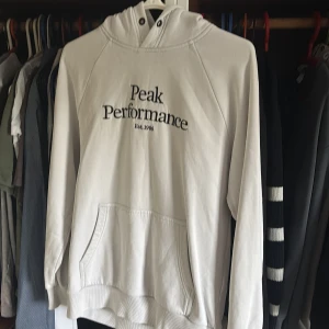 Vit hoodie från Peak Performance - Säljer en stilren vit hoodie från Peak Performance med svart broderad logga på bröstet. Tröjan har huva med snörning, känguruficka och ribbade muddar. Perfekt för dig som gillar en clean och sportig look.