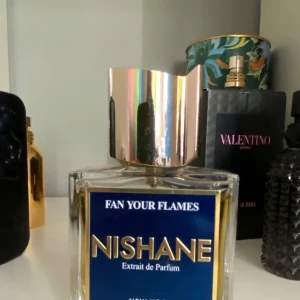 Fan Your Flames Extrait de Parfum från Nishane - Exklusiv parfymflaska från Nishane med doften Fan Your Flames. Flaskan är fyrkantig i genomskinligt glas med en stor, spegelblank silverfärgad kork och blå etikett med guldtext. Lyxig och modern design.