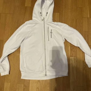 Vit hoodie från Sail Racing med dragkedja - Vit hoodie från Sail Racing med hel dragkedja och extra bröstficka med dragkedja. Broderad logga på bröstet och huva med snygg detalj. Mjuk insida och ribbade muddar. Perfekt för chill eller när du vill ha en clean look.