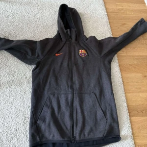 FC Barcelona Nike hoodie - Mörkgrå hoodiejacka från Nike med FC Barcelona-logga på bröstet och orange swoosh. Jackan har dragkedja hela vägen, huva och två stora fickor framtill. Perfekt för dig som älskar fotboll och vill ha en sportig look.