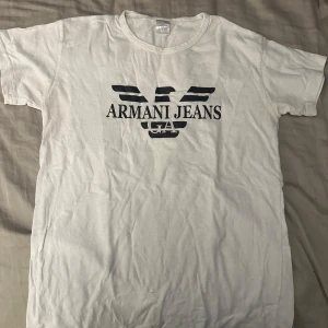 Vit t-shirt från Armani Jeans - Vit t-shirt från Armani Jeans med klassisk svart logga och tryck framtill. T-shirten har rund halsringning och korta ärmar. Perfekt för dig som gillar stilrena och ikoniska plagg med streetkänsla.