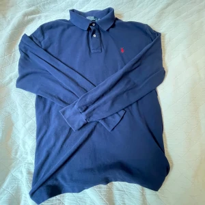 Marinblå pikétröja från Ralph Lauren - Klassisk marinblå långärmad pikétröja från Polo Ralph Lauren med röd broderad logga på bröstet. Tröjan har krage, knappar vid halsen och är tillverkad i mjuk bomull. Perfekt för en stilren och avslappnad look.