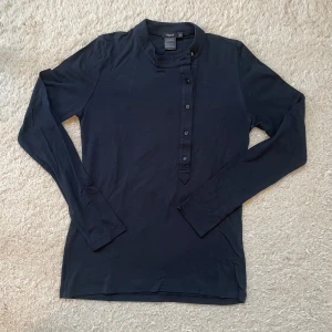 Blå långärmad tröja Filippa K XS - Stilren blå långärmad tröja från Filippa K i mjuk viskosblandning. Tröjan har ståkrage och knappar framtill som ger en clean look. Perfekt för dig som gillar minimalistisk stil och vill ha något som funkar till många outfits.