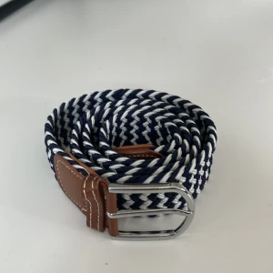 Blåvitt flätat bälte med läderdetaljer - Snyggt flätat bälte i marinblått och vitt med bruna läderdetaljer vid spännet. Bältet har ett klassiskt silverfärgat spänne och är gjort i ett mjukt, flexibelt material som ger en sportig och fräsch look. Perfekt för att lyfta vilken outfit som helst.