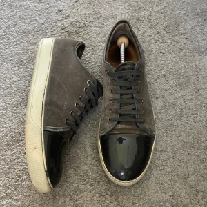 Grå sneakers med svart lackad tå från Lanvin - Säljer ett par grå sneakers från Lanvin med svart lackad tå och vita sulor. Skorna har snörning och en klassisk rund tå. Ovandelen är i mocka och tån är i glansigt material. Perfekta för dig som gillar stilrena detaljer. Inget og kommer med tyvär.