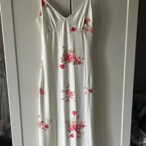 Vit blommig långklänning med smala axelband - Säljer en vit långklänning med rosa blommönster och smala axelband. Klänningen har en figurnära passform och v-ringad halsringning. Perfekt för sommarens alla tillfällen!