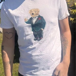 Vit t-shirt med Polo Bear från Ralph Lauren - Vit t-shirt från Ralph Lauren med Polo Bear-tryck på bröstet. Björnen är klädd i smoking och håller ett glas. T-shirten har rund hals och korta ärmar. Slim fit-modell i mjuk bomull.