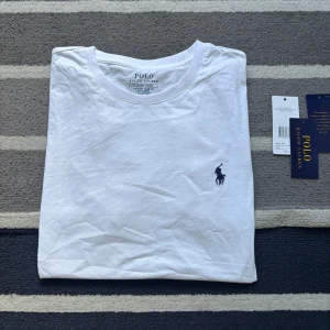 Vit t-shirt från Polo Ralph Lauren - Vit t-shirt från Polo Ralph Lauren med klassisk rund hals och broderad marinblå logga på bröstet. Modellen är custom slim fit och tillverkad i mjuk bomull. Perfekt basplagg med stilren design.