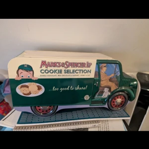 Marks & Spencer Ltd Cookie Selection - En charmig plåtask formad som en retro lastbil från Marks & Spencer, dekorerad med illustrationer av förare och kakor. Perfekt för dig som gillar vintage och samlarobjekt! Asken är tom och passar utmärkt som dekoration eller förvaring.
