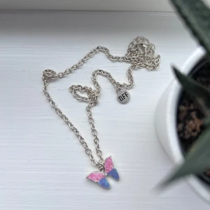 Silverfärgat halsband med fjäril och BFF-berlock - Säljer ett silverfärgat halsband med en söt fjäril i rosa och blått med glittriga detaljer. Halsbandet har även en rund berlock med texten 'BFF'. Perfekt som vänskapssmycke eller present. Önskar du veta exakta mått? Tveka inte att höra av dig!