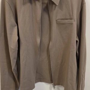 Beige overshirt från Jack & Jones - Säljer en beige overshirt från Jack & Jones med dragkedja framtill och en bröstficka. Jackan har klassisk krage och lång ärm, perfekt för lager-på-lager. Enkel och stilren design som passar till mycket. Nypris 699 kr