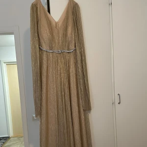 Champagnefärgad långklänning med glitter och bälte - Säljer en elegant champagnefärgad långklänning med glittrigt tyg och långärmade ärmar. Klänningen har v-ringning och ett dekorativt bälte i midjan. Perfekt för bal eller festliga tillfällen.