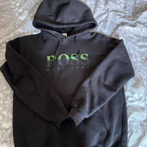 Svart hoodie från Hugo Boss - Säljer en sällsynt svart hoodie från Hugo Boss med stor grön BOSS-logga på bröstet. Tröjan har huva med dragsko och en klassisk magficka. Perfekt för en avslappnad och stilren look.