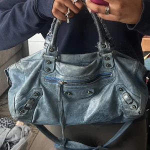 Balenciaga classic city bag - Säljer en äkta Balenciaga Classic City Small i en fantastisk blå färg. Det här är den mindre versionen av den ikoniska City-väskan – perfekt för vardagsbruk men ändå rymlig nog för dina viktigaste saker.  Mått (ca): Bredd: 38 cm Höjd: 24 cm Djup: 14 cm  Ingår: justerbar och avtagbar axelrem Äkta lammnappa med den klassiska mjuka och slitna looken  Silverfärgade metalldetaljer  100 % autentisk  Väskan är i gott använt skick och fullt funktionell.  Reva i ett av handta men det påverkar inte funkti