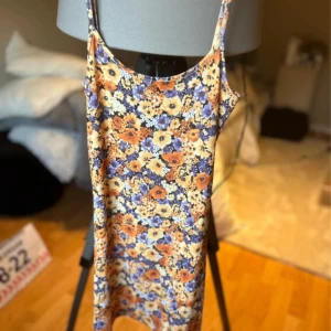 Blommig kortklänning från H&M - Säljer en söt blommig kortklänning från Divided i färgerna blå, orange och beige. Klänningen har smala axelband och är ärmlös med en figurnära passform. Perfekt för varma dagar och enkel att styla med sneakers eller sandaler.🩷