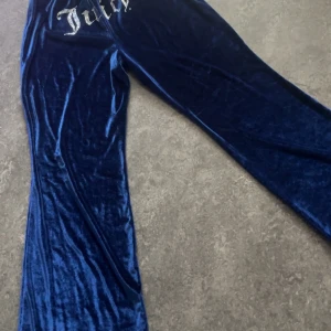 Mörkblå velourbyxor från Juicy Couture - Mörkblå velourbyxor från Juicy Couture med bred resår i midjan och dragsko. Byxorna har raka, vida ben och klassisk Juicy Couture-logga framtill i silver. Perfekta för en avslappnad och trendig look.