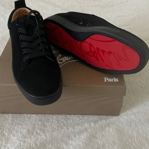 Svarta Louboutin utan spikar  - Säljer ett par svarta sneakers från Christian Louboutin med ikonisk röd sula och svart snörning. Skorna har rund tå och är tillverkade i mocka med läderdetaljer invändigt. Perfekta för dig som vill sticka ut med exklusiv design.