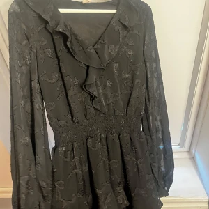 Svart klänning med volang från Nelly - Snygg svart klänning från Nelly med volangdetaljer och långärmad design. Klänningen har markerad midja med smock och subtilt blommönster i tyget. Perfekt för dig som gillar en feminin och trendig look.