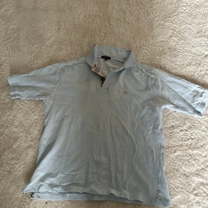 Ljusblå pikétröja från Burberry - Säljer en ljusblå pikétröja från Burberry med klassisk krage och korta ärmar. Tröjan har Burberrys ikoniska rutiga detalj vid knäppningen och är tillverkad i 100% bomull. Perfekt för en stilren och avslappnad look.