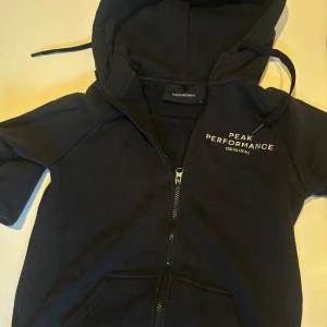 Marinblå hoodie från Peak Performance - Säljer en marinblå hoodie från Peak Performance med dragkedja och vit logga på bröstet. Tröjan har huva med dragsko och fickor framtill. Perfekt för en avslappnad stil.