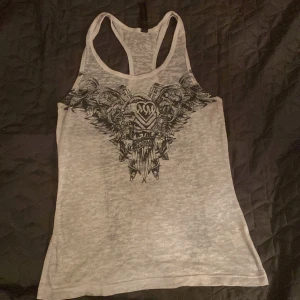 Vit linnetopp med metaltryck - Cool vit linnetopp med svart metalinspirerat tryck både fram och bak. Trycket har detaljer som dödskallar, vingar och texten 'Metal Mulisha'. Modellen är ärmlös med brottarrygg och lätt transparent tyg. Perfekt för dig som gillar edgy och rockig stil.