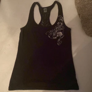 Svart linne - Cool svart linnetopp från metal mulisha med äglavingar och unikt vitt tryck fram och bak. Framsidan har ett detaljerat mönster med text och bakstycket har ett stort vingmotiv. Perfekt för dig som gillar edgy och snygga toppar.