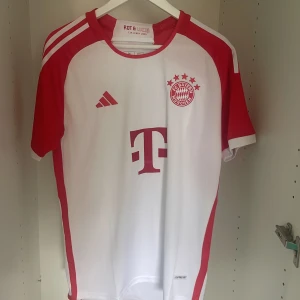 Bayern München Adidas fotbollströja - Snygg Bayern München fotbollströja från Adidas i vitt och rött. Klassisk design med klubbmärke och sponsorlogga på bröstet, samt FC Bayern München-tryck på ryggen. Tröjan är kortärmad och tillverkad i lätt och ventilerande material.