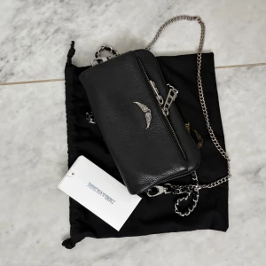 Svart Zadig & Voltaire axelväska i skinn - Snygg svart axelväska från Zadig & Voltaire i äkta skinn med silverfärgad kedjerem och coolt emblem framtill. Äkhetsbevis + Dustbag!! Nästan oanvänd och säljes pga den inte har kommit till användning🔥