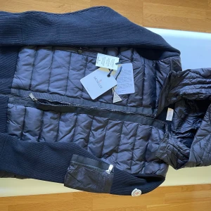 Svart quiltad jacka med huva från Moncler - Säljer en svart quiltad jacka från Moncler med huva och dragkedja. Jackan har ribbade ärmar och detaljer samt en ficka med dragkedja på ena ärmen. Perfekt för kyliga dagar och snygg till vardags. NFC Scan funkar perfekt och det är inget fel med den, helt ny! Den är i storlek 2 alltså m. 