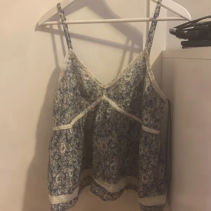 Blommig blå linnetopp med spetsdetaljer - Supersöt blå linnetopp med vitt blommönster och fina spetsdetaljer. Toppen har smala axelband och en lös passform, perfekt för varma dagar. Lätt och luftig känsla med romantisk touch. Den är i storlek s men funkar som m 
